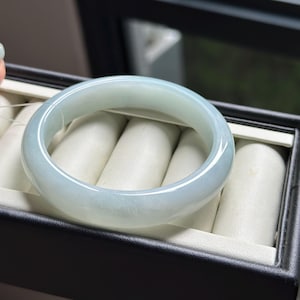 Pode incluir: Um bracelete de jade verde-p&aacute;lido, transl&uacute;cido, polido e liso, com um perfil arredondado. A pe&ccedil;a de joalharia circular apresenta varia&ccedil;&otilde;es subtis na cor e na claridade, exibindo um aspeto suave e gelado em toda a pedra.