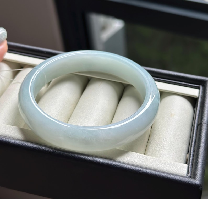 Pode incluir: Um bracelete de jade verde-p&aacute;lido, transl&uacute;cido, polido e liso, com um perfil arredondado. A pe&ccedil;a de joalharia circular apresenta varia&ccedil;&otilde;es subtis na cor e na claridade, exibindo um aspeto suave e gelado em toda a pedra.