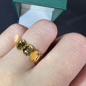 May include: A gold ring with a series of overlapping, textured circular elements. The ring is displayed on a finger, with a green jewellery box in the background. The ring's design gives it a unique, organic appearance.