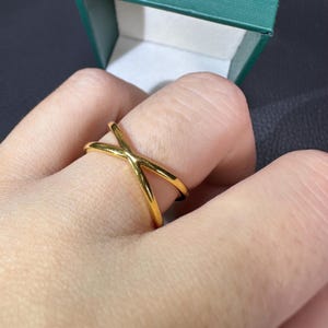 May include: A gold ring with a crisscross design. The ring is made of a shiny, reflective metal and is worn on a finger. The ring's design features two bands that intersect to form an X shape. The ring is a piece of jewellery.