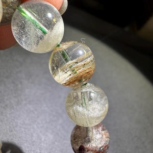 May include: A strand of spherical beads made of clear quartz with internal inclusions. The beads feature visible green needle-like structures, brown fibrous mineral deposits, and cloudy white patches suspended within the transparent stone material.