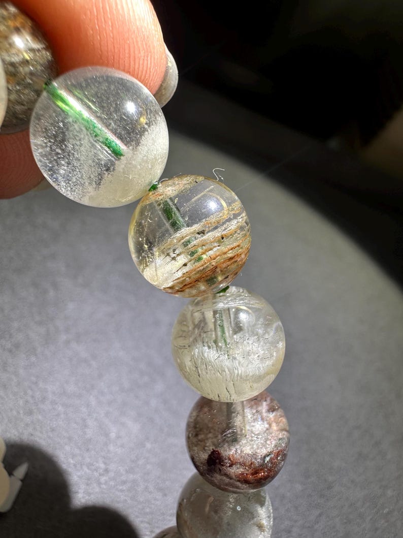 May include: A strand of spherical beads made of clear quartz with internal inclusions. The beads feature visible green needle-like structures, brown fibrous mineral deposits, and cloudy white patches suspended within the transparent stone material.