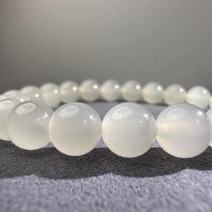 K&ouml;nnte beinhalten: Ein Armband bestehend aus glatten, runden, durchscheinenden wei&szlig;en Perlen. Die Perlen haben eine polierte, gl&auml;nzende Oberfl&auml;che, die das Licht reflektiert und einen sanften Schimmer auf jeder einzelnen Kugel erzeugt, die in einem Kreis angeordnet ist.