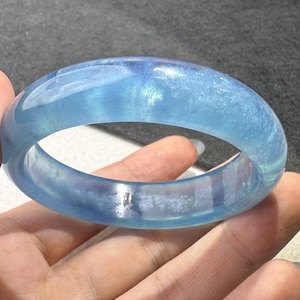 May include: A translucent, light blue bangle bracelet with a smooth, polished surface. The circular accessory features subtle internal variations and a glossy finish that reflects light, showcasing a clear, cool-toned aesthetic throughout the piece.