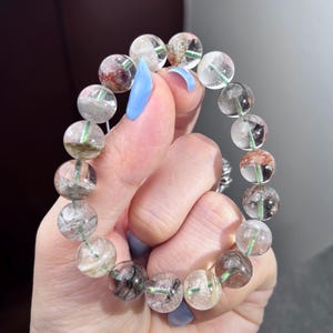 May include: A beaded bracelet composed of round, translucent quartz beads. Each bead contains unique natural inclusions, including dark green, black, and reddish-brown mineral deposits visible within the clear, polished stone spheres.