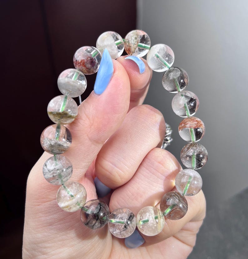 May include: A beaded bracelet composed of round, translucent quartz beads. Each bead contains unique natural inclusions, including dark green, black, and reddish-brown mineral deposits visible within the clear, polished stone spheres.