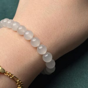 K&ouml;nnte beinhalten: Ein elastisches Armband bestehend aus glatten, runden, halbtransparenten wei&szlig;en Perlen mit sanftem Glanz. Die Perlen sind einheitlich gro&szlig; und auf einem einzelnen Strang aufgereiht. Ein separates Armband aus goldfarbener Metallkette ist daneben sichtbar.