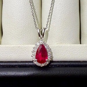 Puede incluir: Un collar de tono plateado con un colgante de piedra preciosa de color rojo intenso en forma de pera, rodeado por un halo de pequeñas piedras claras y brillantes. El colgante cuelga de una cadena fina y delicada con acabado pulido.