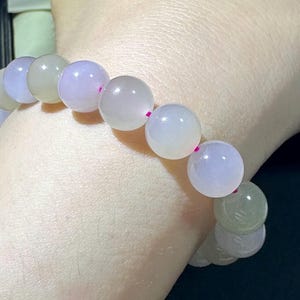 Pode incluir: Uma pulseira de contas com pedras polidas, redondas e lisas em tons variados de lavanda translúcido, roxo pálido e verde sálvia suave. As contas estão enfiadas num cordão fino de cor rosa brilhante que é visível entre cada pedra individual.