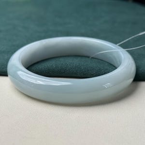 Pode incluir: Uma pulseira r&iacute;gida de jade em tom cinza-esverdeado claro, polida e lisa, com um perfil arredondado e apar&ecirc;ncia transl&uacute;cida. Esta pe&ccedil;a de joalharia circular apresenta um acabamento brilhante e uma espessura constante em toda a circunfer&ecirc;ncia.