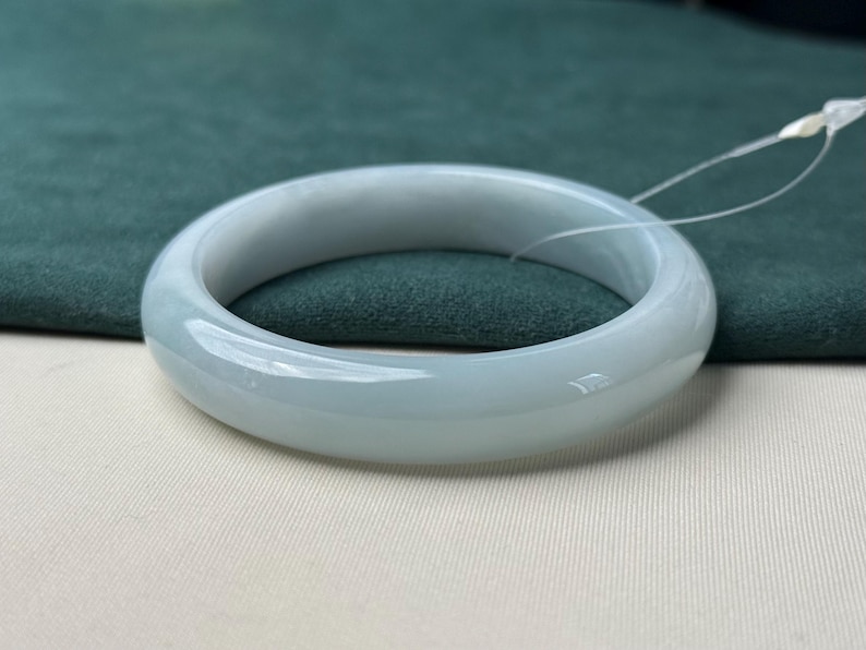 Pode incluir: Uma pulseira r&iacute;gida de jade em tom cinza-esverdeado claro, polida e lisa, com um perfil arredondado e apar&ecirc;ncia transl&uacute;cida. Esta pe&ccedil;a de joalharia circular apresenta um acabamento brilhante e uma espessura constante em toda a circunfer&ecirc;ncia.