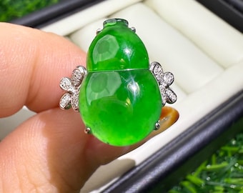 May include: A vibrant green jade ring in the shape of a gourd, set with a silver band and adorned with diamond-studded floral accents. The jade has a glossy finish, and the ring is held up by a hand.