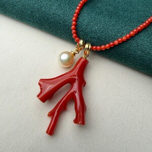 Op de afbeelding: Een rode koraal hangerketting met een parel en gouden accenten. De ketting heeft een felrode koraaltak hanger en een streng kleine rode kralen. De parel heeft een cr&egrave;mewitte kleur. De ketting wordt gepresenteerd op een groene en witte achtergrond.