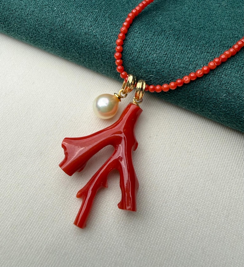 Op de afbeelding: Een rode koraal hangerketting met een parel en gouden accenten. De ketting heeft een felrode koraaltak hanger en een streng kleine rode kralen. De parel heeft een cr&egrave;mewitte kleur. De ketting wordt gepresenteerd op een groene en witte achtergrond.
