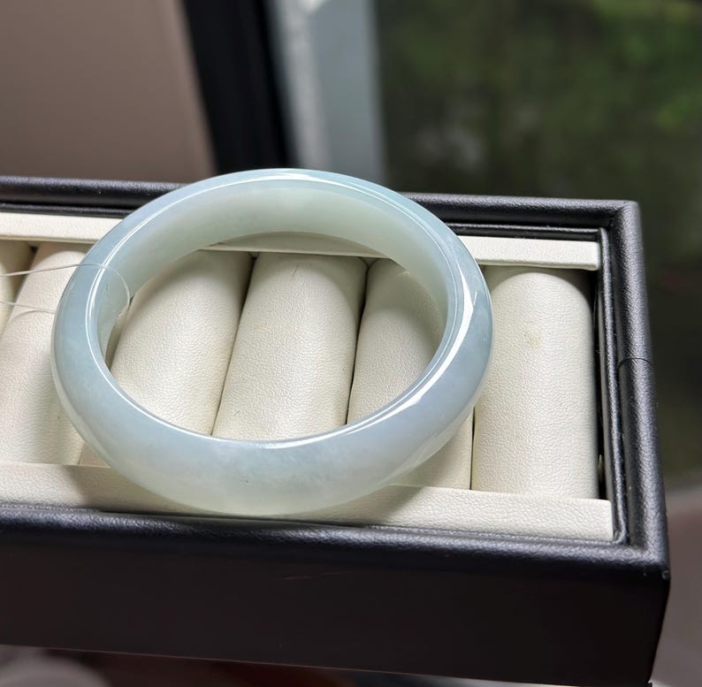 Pode incluir: Uma pulseira tipo bracelete de jade verde p&aacute;lido transl&uacute;cido, polida e lisa, com um perfil arredondado. A pe&ccedil;a de joalharia circular apresenta varia&ccedil;&otilde;es subtis de cor e clareza, refletindo a luz na sua superf&iacute;cie curva e brilhante.