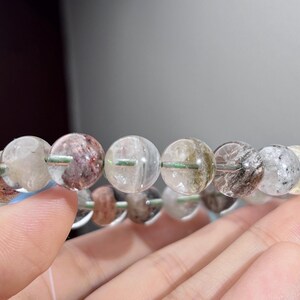 May include: A beaded bracelet composed of round, translucent quartz beads. Each bead features unique internal mineral inclusions in shades of red, brown, black, and white, creating a varied, natural appearance throughout the circular strand.