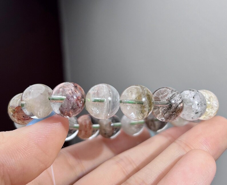 May include: A beaded bracelet composed of round, translucent quartz beads. Each bead features unique internal mineral inclusions in shades of red, brown, black, and white, creating a varied, natural appearance throughout the circular strand.