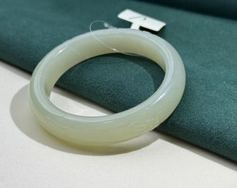 May include: A pale green jade bangle bracelet. The smooth, polished surface of the bracelet has subtle floral carvings. The bracelet is displayed on a dark green fabric, with a white tag attached.
