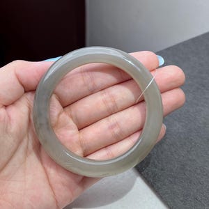 May include: A smooth, translucent grey bangle bracelet with a polished finish. The circular band features subtle, faint carved details along its surface, showcasing a cool-toned, semi-transparent appearance with soft variations in the grey color.
