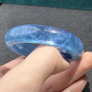 Puede incluir: Un brazalete rígido de color azul translúcido con una superficie lisa y pulida. El material presenta estructuras cristalinas internas e inclusiones que reflejan la luz a lo largo de la banda circular, creando una profundidad de color variada.