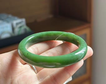 May include: A smooth, polished, circular bangle bracelet crafted from solid green jade. The material features a translucent quality with subtle variations in color intensity and natural dark inclusions throughout the rounded, continuous band.