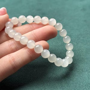 K&ouml;nnte beinhalten: Ein elastisches Armband bestehend aus glatten, runden, halbtransparenten wei&szlig;en Mondsteinperlen. Die Perlen haben eine polierte Oberfl&auml;che und sind in einem kreisf&ouml;rmigen Muster aufgereiht, wodurch eine durchgehende Schleife aus milchig-wei&szlig;en Kugeln entsteht.