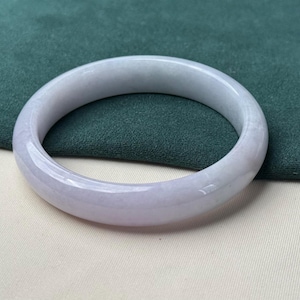 Könnte beinhalten: Ein Armreif aus Lavendel-Jade. Das glatte, polierte Armband hat eine abgerundete Form und einen subtilen Farbverlauf, der von einem blassen Lavendel zu einem sanften Weiß übergeht. Der Jade-Armreif liegt vor einem grünen und weißen Hintergrund.
