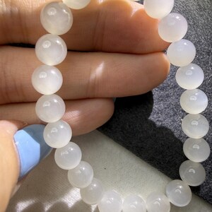 K&ouml;nnte beinhalten: Ein Perlenarmband aus glatten, runden, durchscheinenden wei&szlig;en Mondsteinperlen. Die Perlen weisen einen weichen, milchigen Glanz und ein subtiles Leuchten auf, das das Licht auf ihren polierten Oberfl&auml;chen in einer kreisf&ouml;rmigen Anordnung reflektiert.