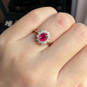 Puede incluir: Un anillo de oro con una piedra preciosa roja vibrante de corte ovalado en el centro, rodeada por un halo de doce pequeñas piedras claras de corte redondo dispuestas en forma de estrella. La banda es fina y metálica.