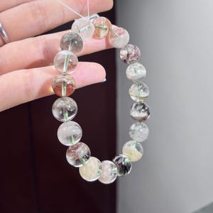 May include: A beaded bracelet composed of round, translucent quartz stones. Each bead features unique internal inclusions in shades of green, red, black, and yellow, creating a varied, multi-colored appearance throughout the circular strand.