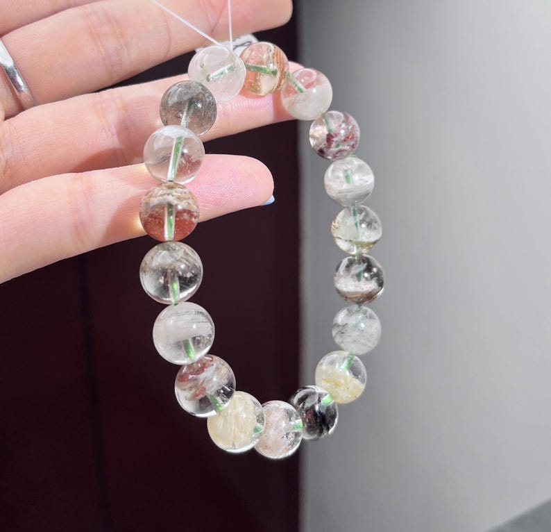May include: A beaded bracelet composed of round, translucent quartz stones. Each bead features unique internal inclusions in shades of green, red, black, and yellow, creating a varied, multi-colored appearance throughout the circular strand.