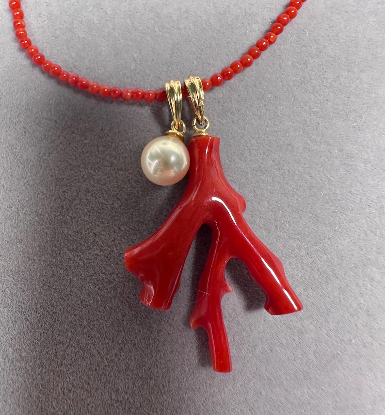 Op de afbeelding: Een rode koraal hanger ketting met een parel en gouden accenten. De ketting heeft een koraal tak hanger en een streng kleine rode kralen. De parel is rond en wit. Gouden accenten verbinden de hanger met de ketting.