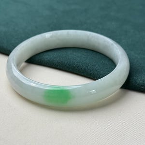 Op de afbeelding: Een jade armband in lichtgroene en witte tinten. De armband heeft een glad, gepolijst oppervlak en een cirkelvorm. De jade heeft een doorschijnende kwaliteit, met een vlek van heldergroene kleur.