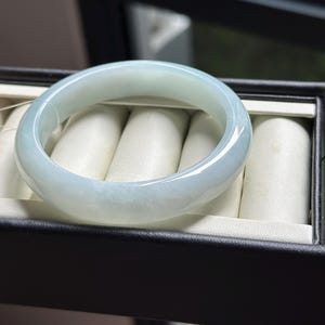 Pode incluir: Uma pulseira tipo bracelete de jade verde-p&aacute;lido, lisa e polida, com apar&ecirc;ncia transl&uacute;cida e perfil arredondado, repousando sobre um suporte de veludo creme dentro de uma caixa de joias retangular preta.
