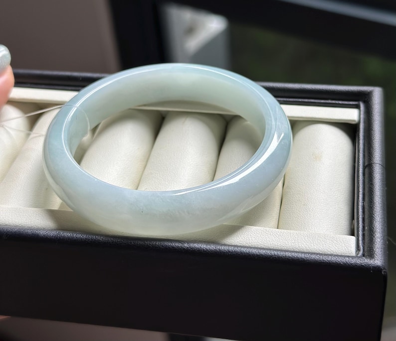 Pode incluir: Uma pulseira tipo bracelete de jade verde-p&aacute;lido, lisa e polida, com apar&ecirc;ncia transl&uacute;cida e perfil arredondado, repousando sobre um suporte de veludo creme dentro de uma caixa de joias retangular preta.