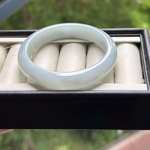 Pode incluir: Uma pulseira de jade verde-p&aacute;lido, lisa e transl&uacute;cida, com um perfil arredondado. A pulseira &eacute; apresentada sobre um suporte de veludo cor de creme para an&eacute;is dentro de uma caixa de joias retangular preta.