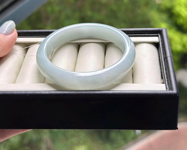 Pode incluir: Uma pulseira de jade verde-p&aacute;lido, lisa e transl&uacute;cida, com um perfil arredondado. A pulseira &eacute; apresentada sobre um suporte de veludo cor de creme para an&eacute;is dentro de uma caixa de joias retangular preta.