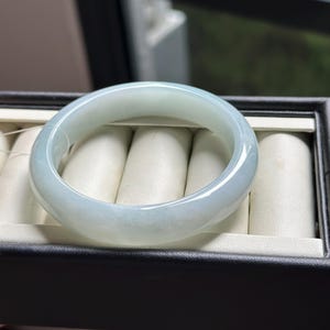 Pode incluir: Um bracelete de jade verde-p&aacute;lido, transl&uacute;cido e liso, com uma superf&iacute;cie polida e arredondada. O bracelete circular apresenta varia&ccedil;&otilde;es subtis na cor e na claridade, refletindo a luz sobre a sua estrutura de pedra s&oacute;lida e curva.