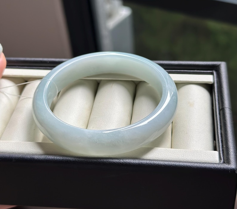 Pode incluir: Um bracelete de jade verde-p&aacute;lido, transl&uacute;cido e liso, com uma superf&iacute;cie polida e arredondada. O bracelete circular apresenta varia&ccedil;&otilde;es subtis na cor e na claridade, refletindo a luz sobre a sua estrutura de pedra s&oacute;lida e curva.