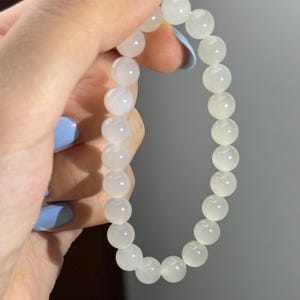 K&ouml;nnte beinhalten: Ein elastisches Armband bestehend aus glatten, runden, durchscheinenden wei&szlig;en Perlen. Die Perlen haben ein subtiles milchiges Aussehen und sind in einer durchgehenden kreisf&ouml;rmigen Schleife aneinandergereiht, was ein schlichtes Design ergibt.