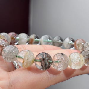 May include: A beaded bracelet featuring round, translucent quartz beads with internal inclusions in shades of black, brown, and white. The beads are strung on a thin, light green cord, creating a circular arrangement of polished, natural stone spheres.