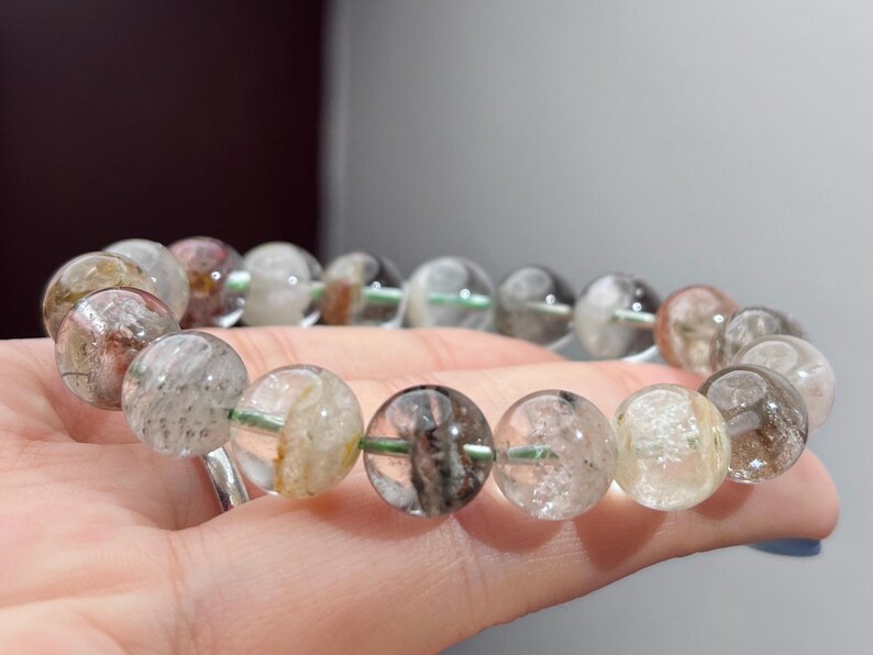 May include: A beaded bracelet featuring round, translucent quartz beads with internal inclusions in shades of black, brown, and white. The beads are strung on a thin, light green cord, creating a circular arrangement of polished, natural stone spheres.