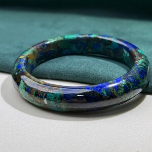 May include: A polished, circular bracelet with a marbled pattern of deep blues, greens, and hints of gold. The gemstone bracelet is set against a teal background, showcasing its natural color variations and glossy finish. A unique piece of jewelry.