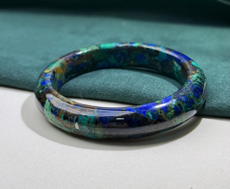 May include: A polished, circular bracelet with a marbled pattern of deep blues, greens, and hints of gold. The gemstone bracelet is set against a teal background, showcasing its natural color variations and glossy finish. A unique piece of jewelry.