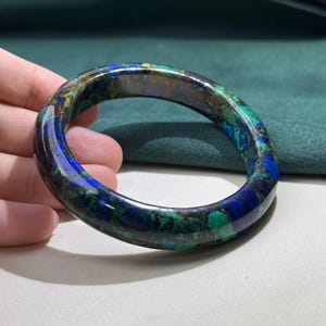 May include: A polished, circular bracelet with a marbled appearance. The bracelet features a blend of deep blues, greens, and browns, creating a natural, gemstone-like aesthetic. The smooth surface reflects light, highlighting the varied colors and patterns.