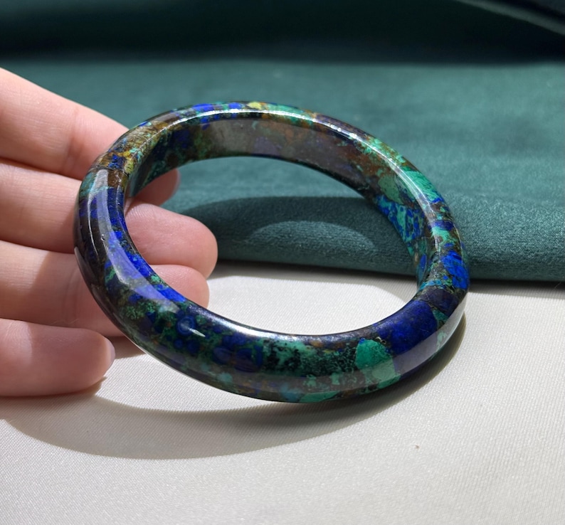 May include: A polished, circular bracelet with a marbled appearance. The bracelet features a blend of deep blues, greens, and browns, creating a natural, gemstone-like aesthetic. The smooth surface reflects light, highlighting the varied colors and patterns.