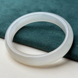 Può includere: Un braccialetto bangle in giada bianco traslucido. Il braccialetto liscio e circolare è esposto su uno sfondo bianco e verde acqua. La giada è di colore pieno e ha una finitura lucida.