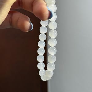 K&ouml;nnte beinhalten: Ein elastisches Armband bestehend aus glatten, runden, halbtransparenten wei&szlig;en Perlen. Die Perlen haben eine polierte, gl&auml;nzende Oberfl&auml;che und sind in einer einzigen durchgehenden Schlaufe aufgereiht, was eine gleichm&auml;&szlig;ige, runde Form ergibt.