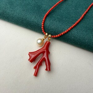 Op de afbeelding: Een rode koraal ketting met een takvormige hanger en een enkele parel. De ketting heeft kleine rode kralen en goudkleurige sluitingen. De hanger is felrood en de parel is cr&egrave;mewit.