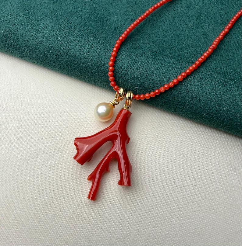 Op de afbeelding: Een rode koraal ketting met een takvormige hanger en een enkele parel. De ketting heeft kleine rode kralen en goudkleurige sluitingen. De hanger is felrood en de parel is cr&egrave;mewit.